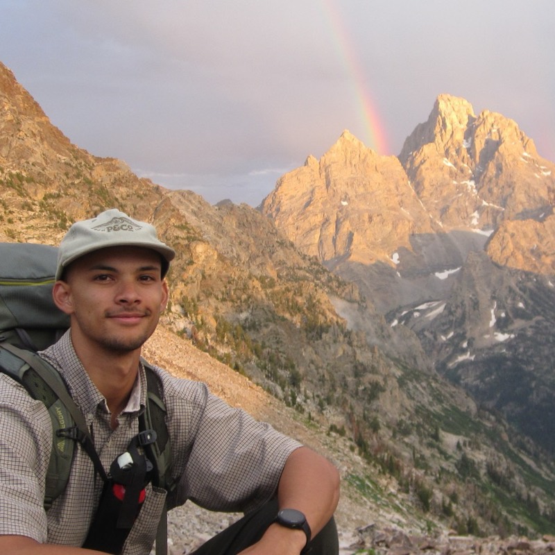 Sorell Miller, owner of Rooted Search Co., smiling in a cap and backpack on a mountain trail with sunlit peaks and a rainbow behind him